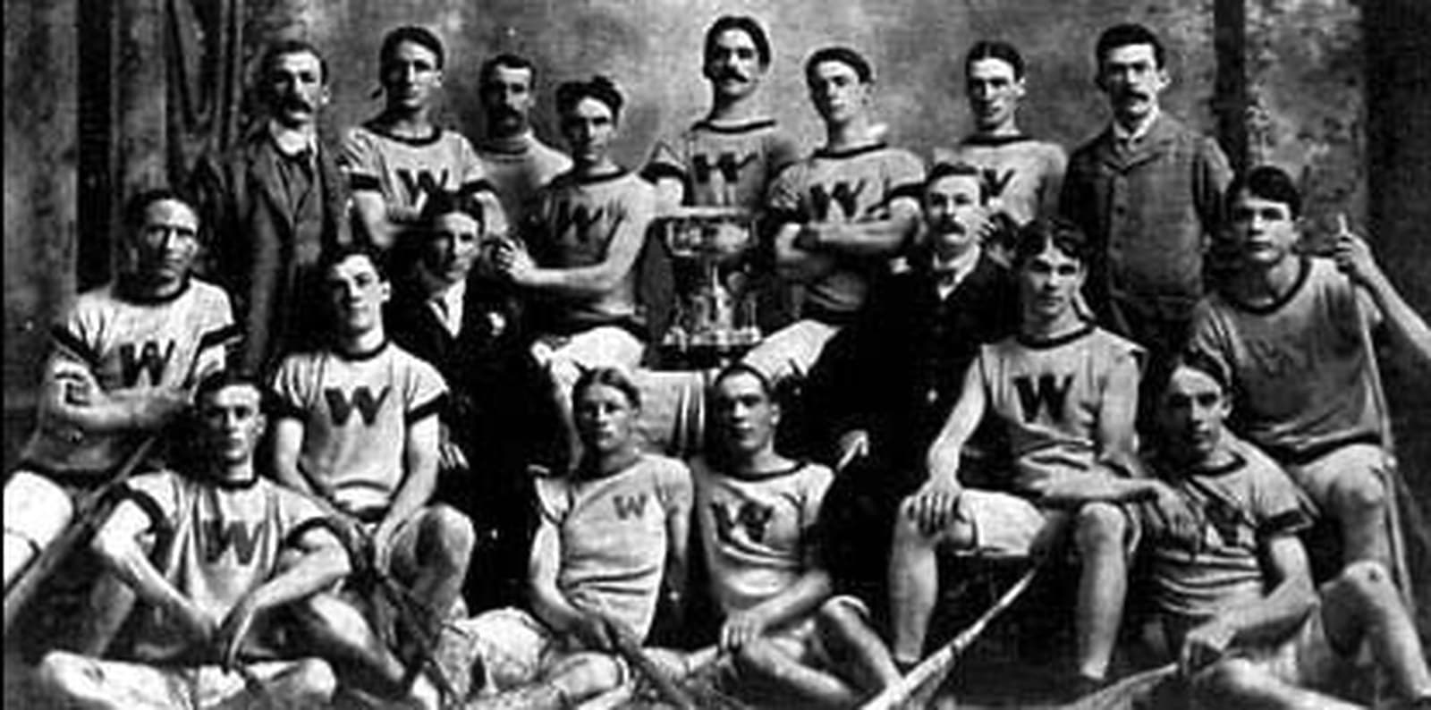 A black-and-white group photograph of about sixteen men. Several in front are seated or kneeling in dark jerseys with a large letter W on the chest, some holding long-handled lacrosse sticks with netted heads. A two-handled trophy cup sits at the centre of the group. Other men, in suits and ties rather than uniforms, stand or sit interspersed among the players.