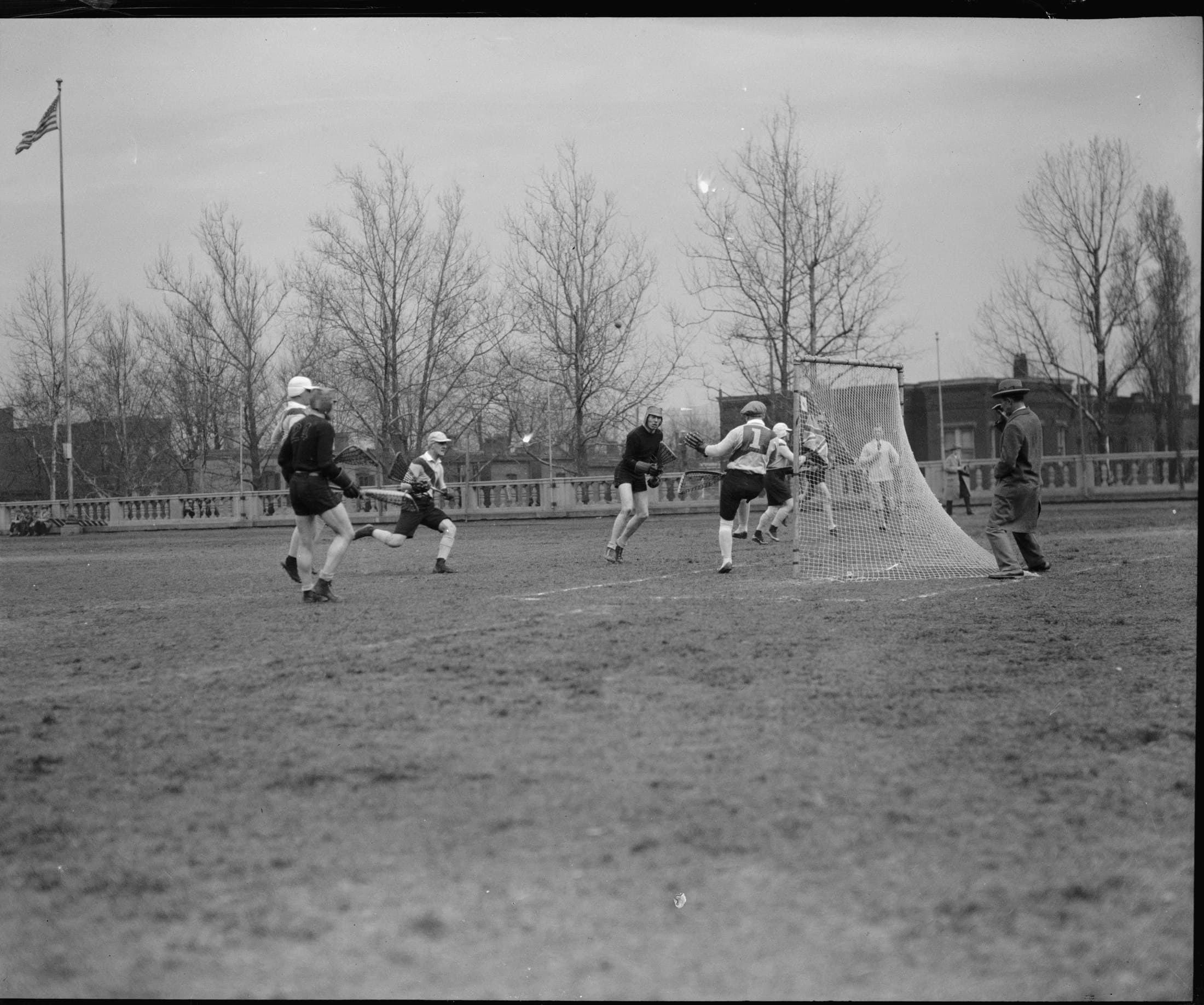 A grainy black-and-white photograph of a lacrosse game in progress on an open field ringed by a low stone balustrade, bare trees and rowhouses behind it. Several players in shorts and jerseys converge on a goal with a netted crease behind it; a man in a suit and hat stands at the goal line watching. An American flag flies from a pole at the far side of the field.