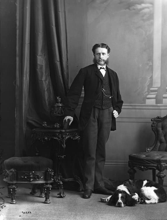 A formal studio portrait of a young man with a full beard and moustache, standing in a dark suit and bow tie beside a small ornamented table, one hand resting on a stack of books. A heavy curtain and a painted backdrop of columns are behind him, and a dog lies on the floor at his feet.