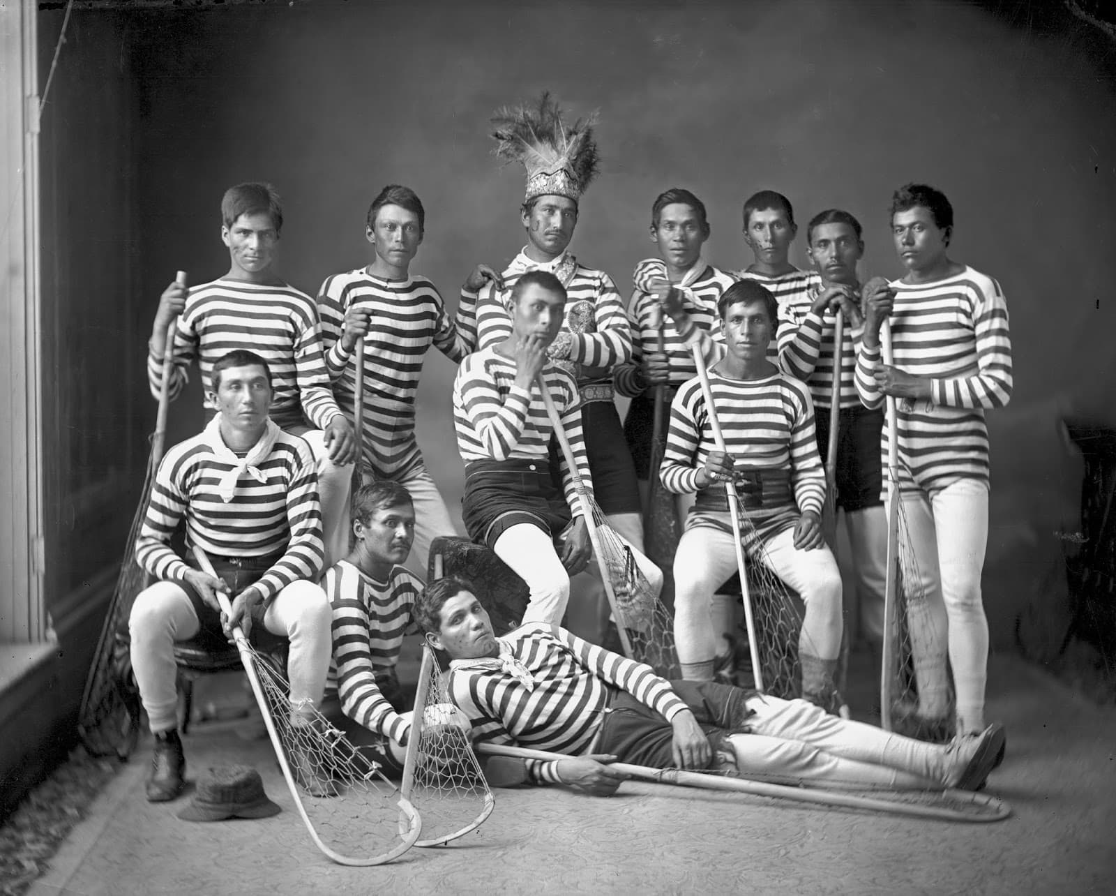 A studio photograph of eleven lacrosse players in matching horizontally striped jerseys, holding wooden sticks with netted heads. Several stand at the back, several sit in front of them, and one lies stretched across the floor at the front. The man standing at the centre wears a tall feathered headdress and an ornamented sash.